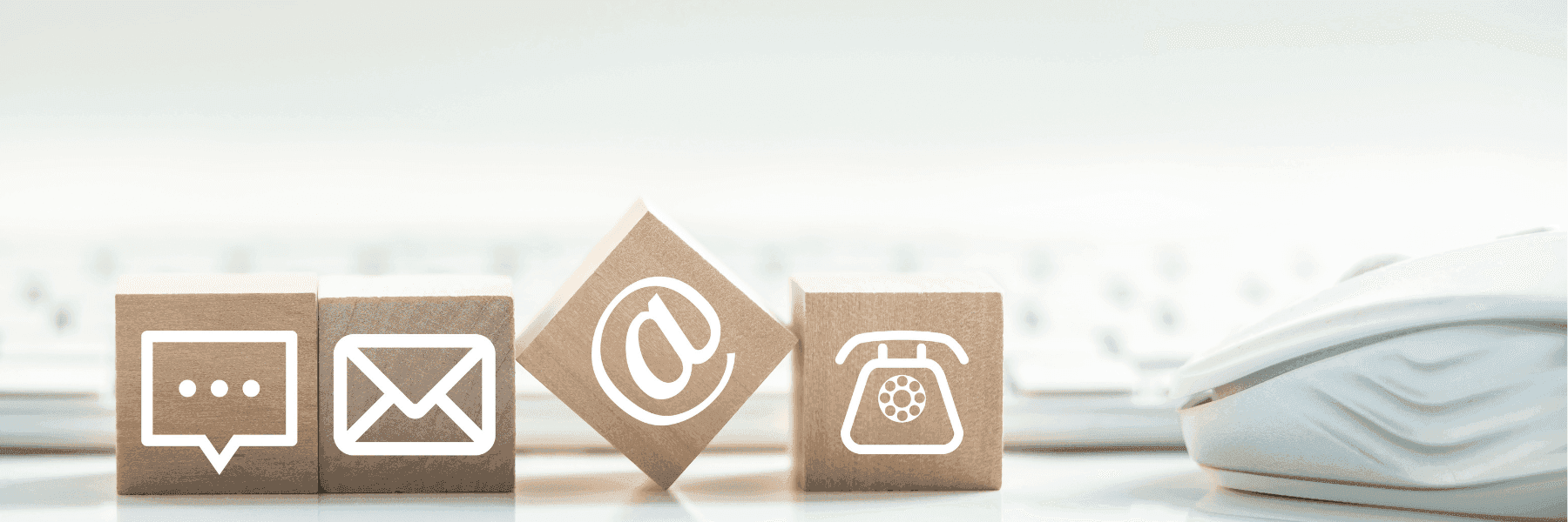 Four wooden blocks displaying contact icons—a speech bubble, envelope, “@” symbol, and rotary phone—sit beside a computer keyboard and mouse.