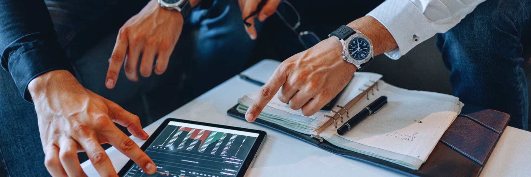 Close-up of business professionals reviewing financial data on a tablet and taking notes in a notebook during an investment meeting.