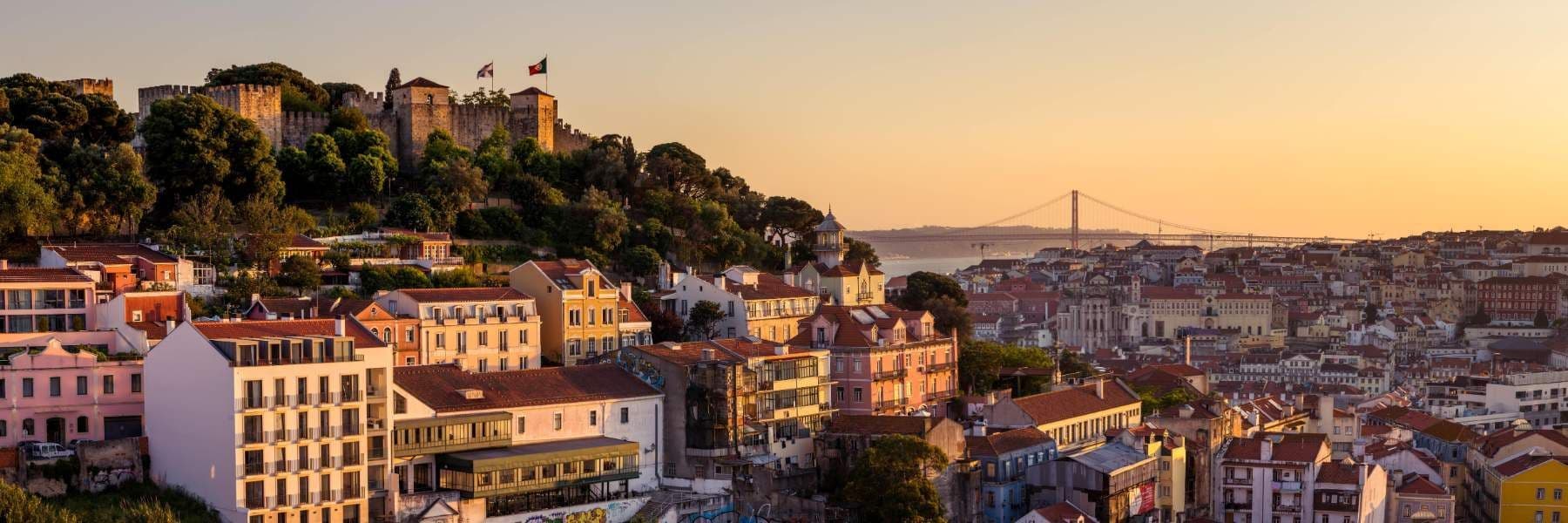 View over Lisbon’s hilly rooftops toward São Jorge Castle perched on a green hill, with the 25 de Abril Bridge spanning the river in the distance at sunset.