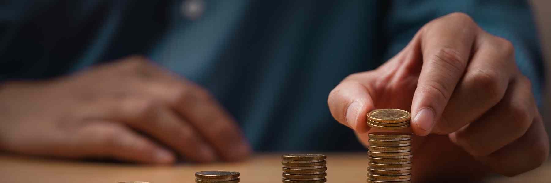 A person’s hand places a coin atop a rising stack beside several smaller piles of coins on a tabletop.