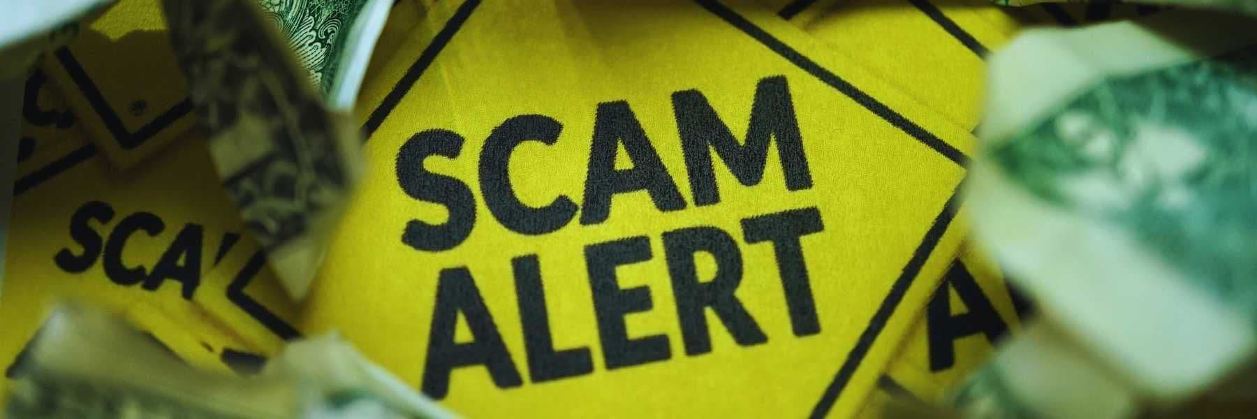 A bright yellow sign with bold black text reading “SCAM ALERT” surrounded by crumpled US dollar bills.