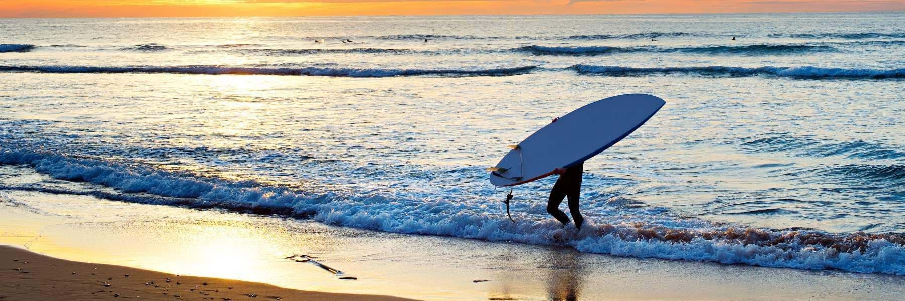 A surfer carrying a surfboard walks into gentle waves on a sandy beach at sunset.