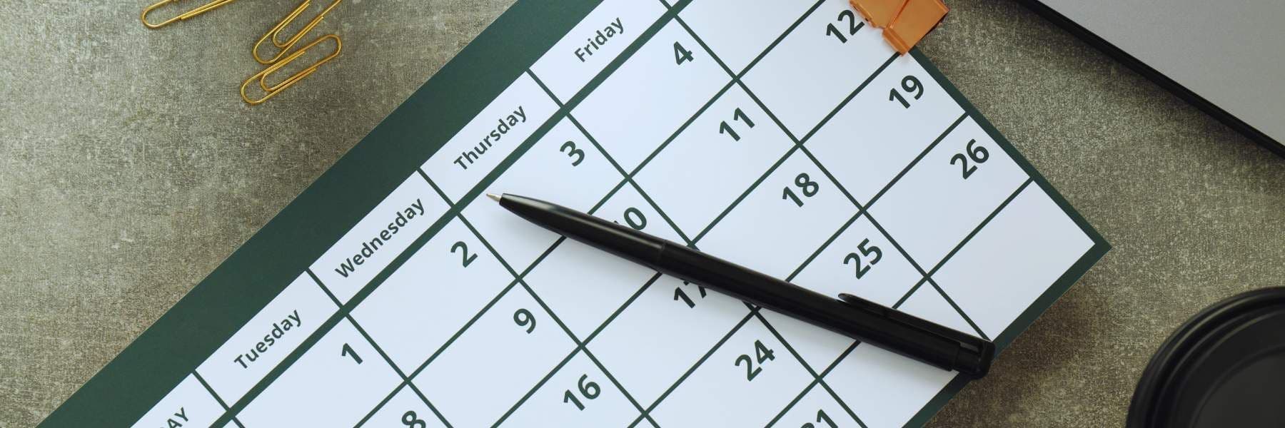 A desk surface featuring a monthly calendar with numbered dates, a black pen resting across it, gold paperclips in the corner, and partial views of a laptop and coffee cup.