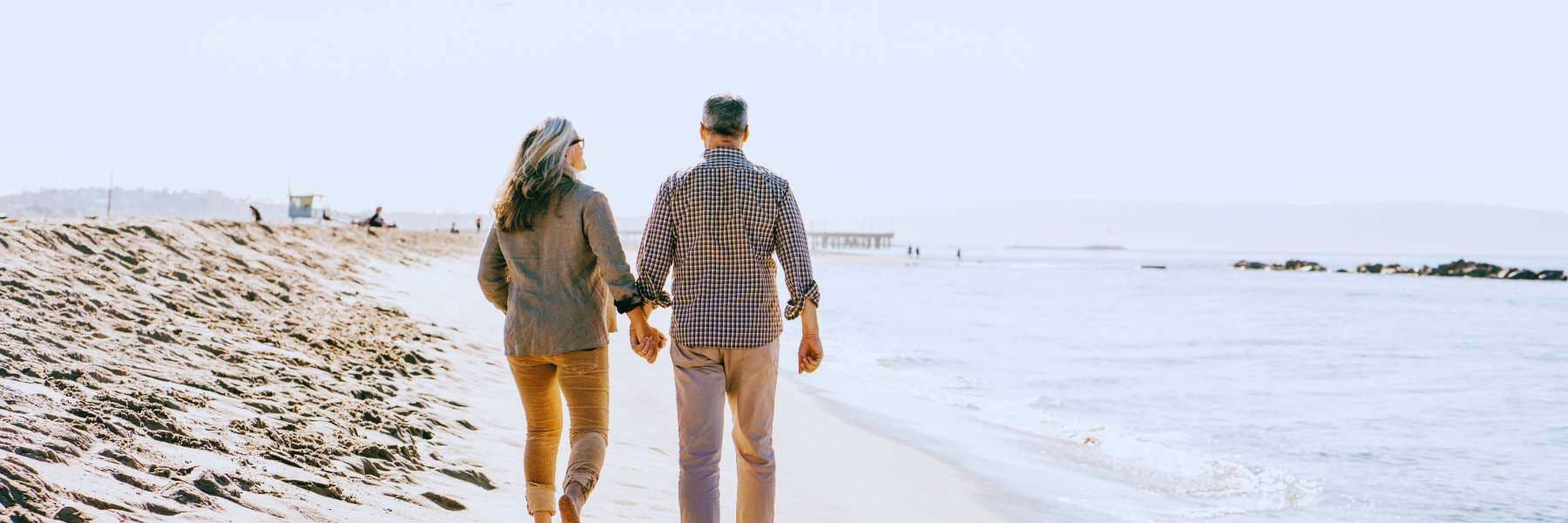 An older couple walks hand in hand along a sandy beach shoreline at dawn.