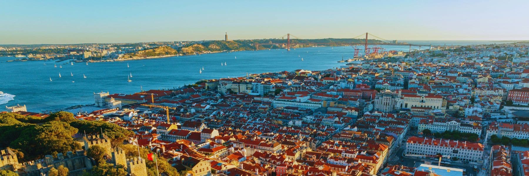A panoramic view of Lisbon, Portugal, featuring the Tagus River, the 25 de Abril Bridge, and the city’s iconic red rooftops at sunset.