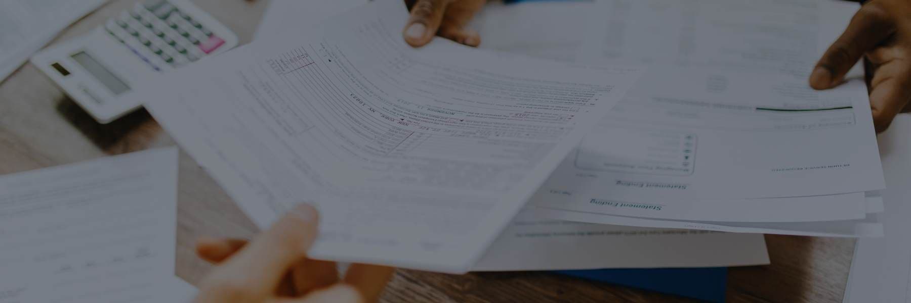 Hands exchanging printed financial documents and forms on a desk, with a calculator visible in the background.