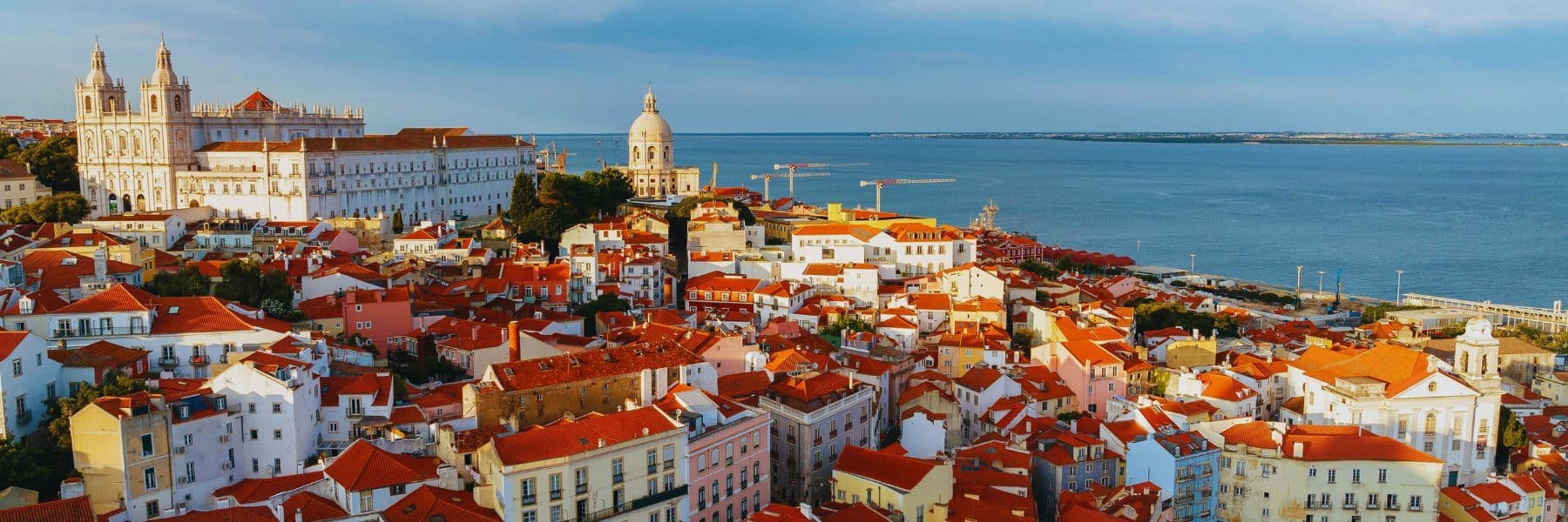 Panoramic view of Lisbon’s Alfama district with red-tiled rooftops, historic churches, and the Tagus River in the background, Portugal.
