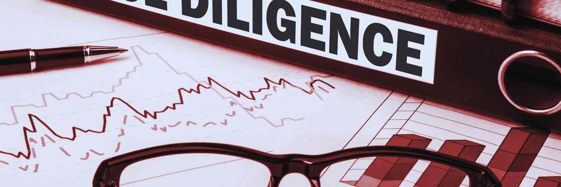 A due diligence binder rests atop financial charts displaying line and bar graphs, with a pen and a pair of glasses alongside.
