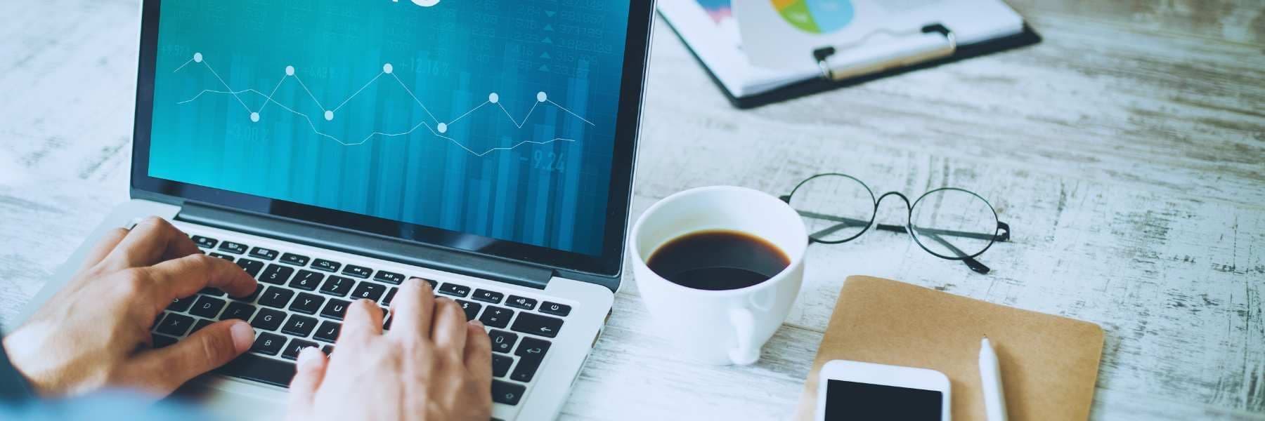 Hands typing on a laptop displaying a line graph, with a coffee cup, eyeglasses, notebook, smartphone, and a clipboard of pie charts arranged on a rustic wooden desk.